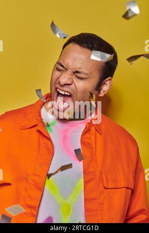 emotional indian man in orange jacket screaming near confetti on yellow backdrop, party Stock Photo