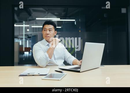 Portrait of a young Asian male businessman in a white shirt working in the office at a laptop. He looks seriously to the side. Stock Photo