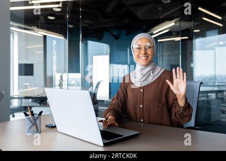 Portrait of a young Muslim woman in hijab sitting at a desk in the office, working and studying on a laptop, looking and waving at the camera. Stock Photo