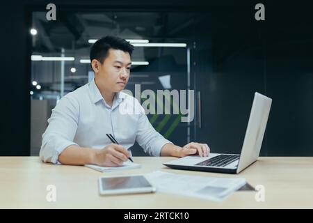 Asian young man accountant, financial analyst, economist is focused in office with laptop and documents. Stock Photo