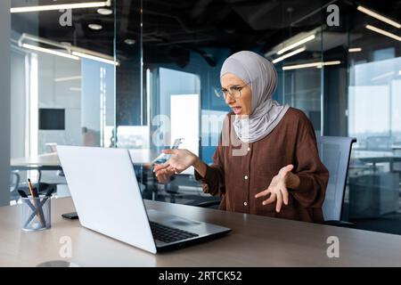 Young muslim woman in hijab is working and studying in office at laptop. He looks upset at the screen and spreads his hands, problems with the Internet, he talks in a video call. Stock Photo
