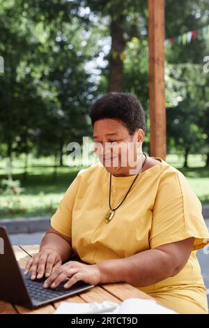Vertical portrait of Black senior woman using laptop outdoors at terrace and smiling looking at computer screen Stock Photo