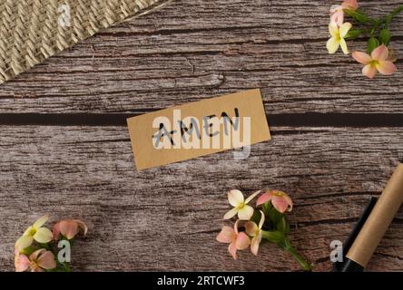 Amen, handwritten text with uppercase letters in English on a wooden table with flowers and pen in vintage style. Top view, flat lay. Stock Photo