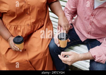 Close up of romantic senior couple holding hands outdoors while sitting on bench in park with coffee, copy space Stock Photo