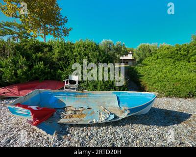 An abandoned and broken boat and the same chair lies on the shore of the Sea, surrounded by bushes and trees with a house in the distance in Greece Stock Photo