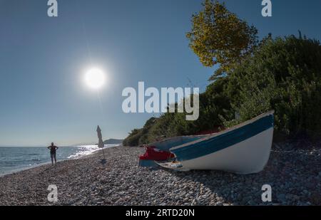 An abandoned and broken boat and the same chair lies on the shore of the Sea, surrounded by bushes and trees with a house in the distance in Greece Stock Photo