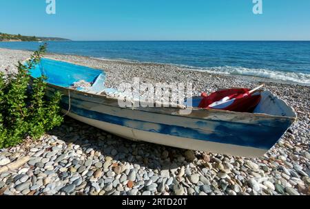 An abandoned and broken boat and the same chair lies on the shore of the Sea, surrounded by bushes and trees with a house in the distance in Greece Stock Photo