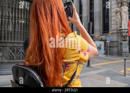 Red-haired girl with straight hair and a yellow dress taking a photo with her mobile phone in the streets of Madrid. Stock Photo