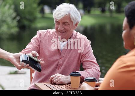 Portrait of smiling senior couple paying via NFC by smartphone at outdoor cafe, copy space Stock Photo