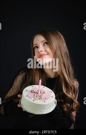 Beautiful happy teen girl wearing black dress holding birthday cake with candle on a plate. Cake with the inscription Hello 14. Girl makes a wish look Stock Photo