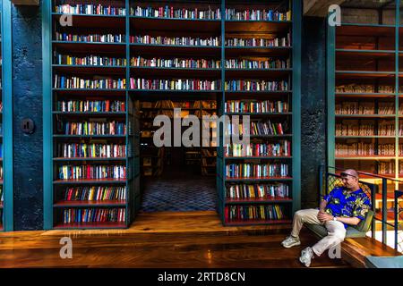 A man sits on a step in the Cafe Stamba library in Tbilisi, Georgia. Bookshelves fill the room. A passageway leads to another room with books Stock Photo