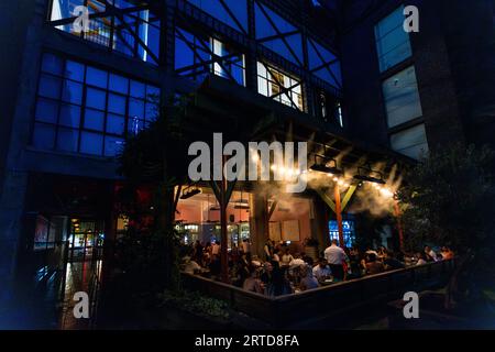 Guests sit outside in the evening at the Cafe Stamba restaurant in Tbilisi, Georgia. A modern building with large windows forms the backdrop. Water is sprayed with a high-pressure system for cooling Stock Photo