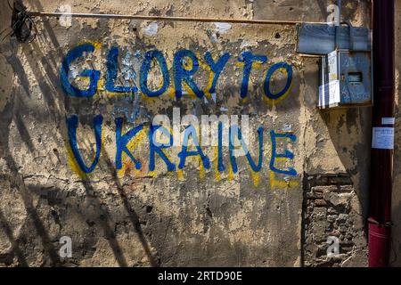 The words "Glory to Ukraine" are painted in blue letters with yellow shadows on a weathered house wall on Arsena Odzelashvili Street in the Mtatsminda district of Tbilisi, Georgia. Blue and yellow are the colours of the Ukrainian flag. The plaster is crumbling, and bricks show through on the right. An electricity box and a red downpipe hang beside the lettering. In 2023, graffiti against Russia and in favour of closer ties with Europe shaped the capital's streetscape. Stock Photo