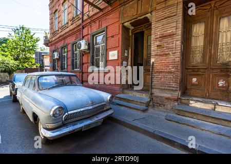 A light blue GAZ-21 Volga is parked in front of a brick house on Arsena Odzelashvili Street in the Mtatsminda district of Tbilisi, Georgia. The car is dusty and scratched, and the chrome of its grille and bumper is intact. The red brick house has barred windows, an open front door and two tall carved wooden doors on the right. A plaque with a Georgian inscription hangs beside the entrance. The Soviet upper-class car was built from 1956 to 1970. In the Soviet Union it was a status symbol. Stock Photo
