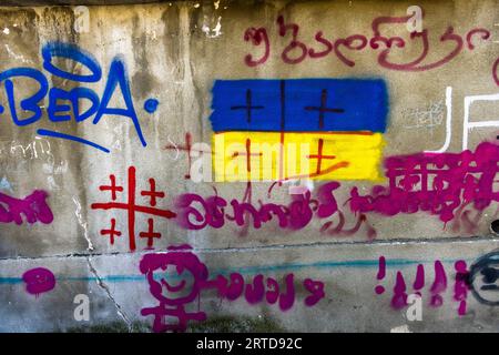 Political graffiti covers a concrete wall on Sulkhan Saba Orbeliani Street in the Sololaki neighbourhood of Tbilisi, Georgia. In the centre is the blue and yellow Ukrainian flag, with red crosses laying the pattern of the Georgian national flag over it. On the left, the red cross motif of the Georgian flag stands on its own. Pink Georgian lettering, a stick-figure face and exclamation marks surround the flags. At top left, the word "BEDA" is written in blue. In 2023, graffiti against Russia and in favour of closer ties with Europe shaped the capital's streetscape. Stock Photo