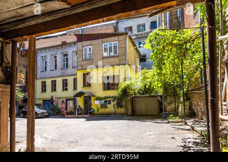A yellow-rendered residential building stands in a backyard in Tbilisi, Georgia, possibly in the Sololaki neighbourhood. The view leads through a covered passageway with wooden beams and supporting posts. On the right, a grapevine climbs over a metal frame. In front of the house, a person sits under a small canopy, with clothes hanging on the façade beside them. A dark car is parked on the left. Taller buildings of brick and grey render rise behind. Stock Photo