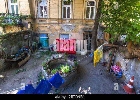 Colourful washing dries on lines in the backyard of an old residential building on Lado Asatiani Street in the Sololaki neighbourhood of Tbilisi, Georgia. In the centre of the yard stands a round masonry enclosure lined with potted plants. A red patterned blanket hangs on the house wall, and pots stand on a wooden rack on the left. The façade shows heavily flaking plaster, and two windows have turquoise and white curtains. A traffic cone leans against the wall on the right. Stock Photo