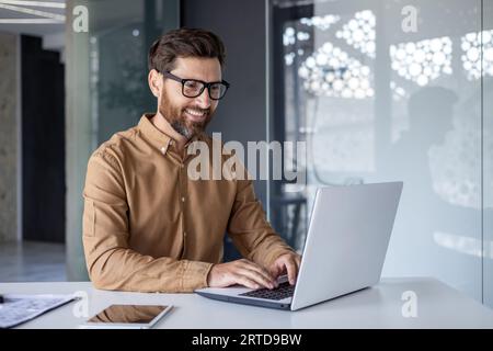 Experienced programmer inside office at workplace, senior man with beard and glasses working on laptop, businessman satisfied with achievement results. Stock Photo