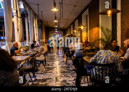 Evening scene on the covered veranda of the Kikodze Coffee & Cocktail Bar in Vardisubani, Tbilisi: guests sit beneath hanging lamps on ornamental concrete tiles, whilst the exposed clinker brick wall and curtains between white columns frame the long restaurant corridor. The bar forms part of a historic complex of buildings in the Vardisubani district. Vasil Petriashvili Street, Vardisubani, Vera, Mtatsminda District, Tbilisi, 0108, Georgia Stock Photo