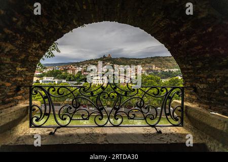 A stone arch with a wrought-iron railing frames the view across the Mtkvari (Kura) River from the Metekhi ascent in the Avlabari district of Tbilisi, Georgia. A riverside road runs along the opposite bank, and residential houses climb a green hillside behind it. A tower stands on the crest as part of Narikala Fortress. Grey clouds hang over the hill. Narikala Fortress was founded in the 4th century. Most of its surviving fortifications date from the 16th and 17th centuries. The capital was completely destroyed 26 times over the course of its history. Stock Photo