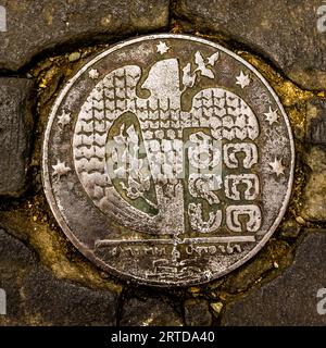 A round metal plaque is set into the cobblestones of Hovanes Tumaniani Street in the Betlemi quarter of the Old Town of Tbilisi, Georgia. It shows a stylised eagle with scale-like wings. Its body bears an oval field with an irregular outline and Georgian letters in an old script. A small bird flies at the upper right. A line with small lettering runs beneath the eagle, with a motif of horizontal lines below it. Stars surround the image along the rim. Green patina has formed in the recesses. Stock Photo