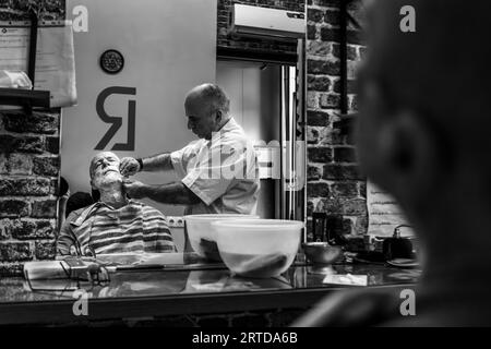 A barber leans over an elderly customer in a barber's shop on Pushkin Street in the Old Town of Tbilisi, Georgia. He holds the customer's lathered face with his hands. The customer reclines with his eyes closed, a striped cape over his shoulders. The scene appears in the salon's mirror, which is why the "RL" logo on the wall is reversed. Two white bowls stand on a glass shelf in front of the mirror. An air conditioner hangs on the brick wall. Stock Photo