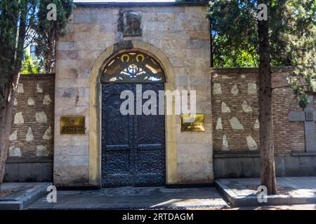 The entrance gate of the Patriarchate of Georgia stands on Erekle II Square in the Old Town of Tbilisi, Georgia. The dark double door bears reliefs of two angels. A round arch of pale ashlar blocks frames it, with an ornamental grille and a cross in the fanlight. An icon relief, possibly of the Mother of God, is set into the wall above the arch. Two gold-coloured plaques beside the gate bear the inscription "Patriarchate of Georgia" in Georgian and English. The Patriarchate heads the Georgian Orthodox Church. A brick wall with inset fieldstones adjoins on both sides. A stone cross stands on th Stock Photo