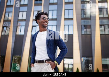 Confident african American male in stylish clothes stands in city next to modern building, looking to side with smile. Happy black man with hands in p Stock Photo
