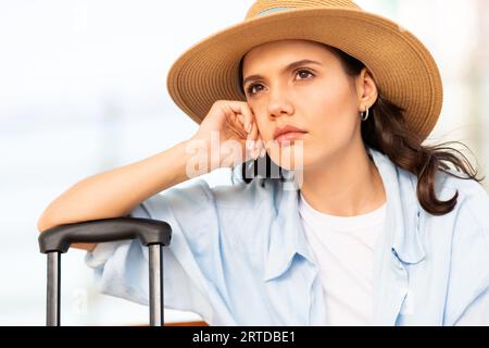 Unhappy sad young caucasian lady in hat with suitcase looks at empty space, suffer from late Stock Photo