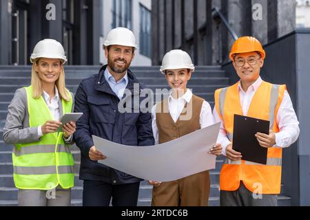 Team of architects builders, diverse group of men and women, inspecting construction stages of office building from outside, holding plan drawing on paper in hands, smiling and looking at camera. Stock Photo