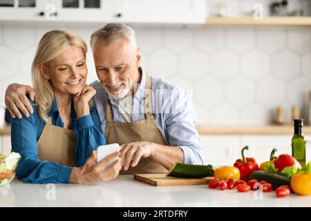 Smiling senior spouses using smartphone in kitchen while cooking food together Stock Photo