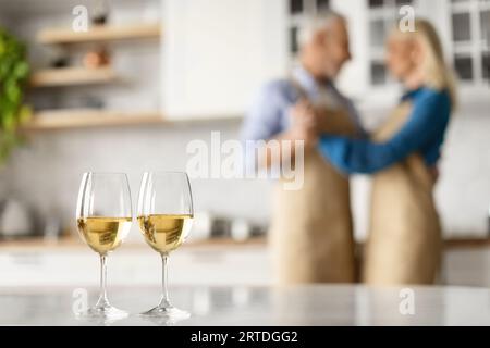 Selective focus on two glasses of white wine on kitchen table over dancing senior couple Stock Photo