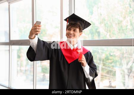 Male graduate student with diploma taking selfie near window in room Stock Photo