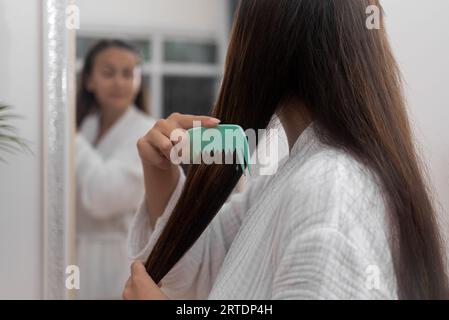 Woman combing long straight chestnut hair looking in mirror in bathroom hairstyling with comb Stock Photo