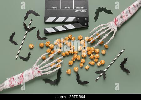 Composition with tasty caramel popcorn, clapperboard, paper bats, skeleton hands and straws for Halloween celebration on green background Stock Photo