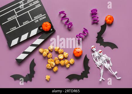 Composition with tasty caramel popcorn, clapperboard, paper bats, skeletons and pumpkins for Halloween celebration on purple background Stock Photo