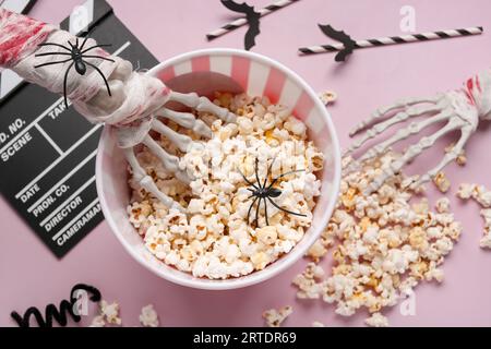 Bucket with tasty popcorn, skeleton hands and clapperboard for Halloween celebration on pink background Stock Photo