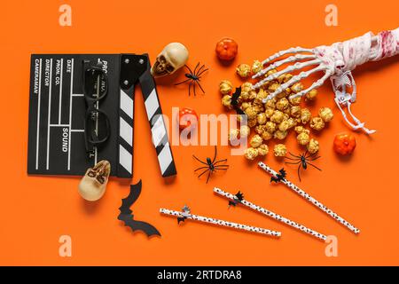 Composition with tasty caramel popcorn, clapperboard, straws, skulls and skeleton hand for Halloween celebration on orange background Stock Photo