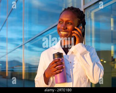 young female doctor talking by phone while holding a coffee thermo in front of a hospital Stock Photo