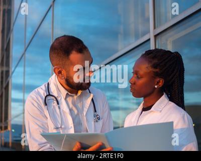 Two hospital doctors, a woman and a man, taking a break outdoors. Looking at each other while smiling. Stock Photo
