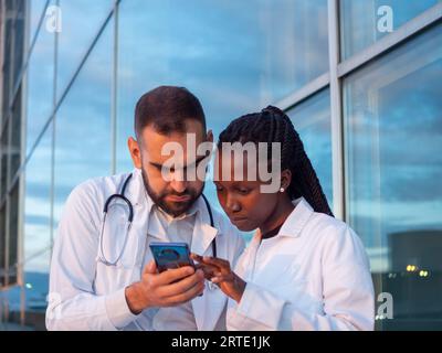 Two hospital doctors, a woman and a man, taking a break outdoors and watching something on their phone Stock Photo