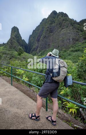 View taken from behind of a man standing on a walkway photographing the view of the Kuka‘emoku, Iao Needle in the lush, Iao Valley in Central Maui Stock Photo