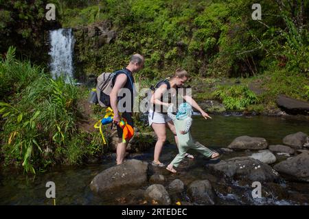 A family carefully walking on a path of volcanic rocks while crossing a stream with a beautiful waterfall in the background on the Road to Hana Stock Photo