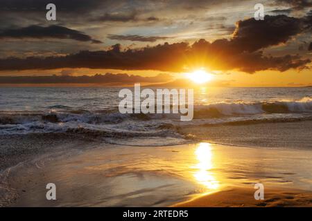 Ocean waves along the shoreline of Kamaole 2 Beach at twilight with a golden sun glowing under a cloudy sky and reflecting on the shore Stock Photo