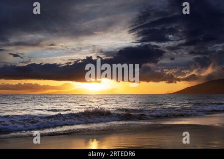 Ocean waves along the shoreline of Kamaole 2 Beach at twilight with a golden sun glowing under a cloudy sky Stock Photo