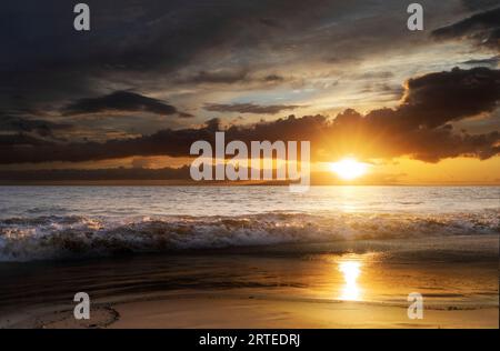 Ocean waves along the shoreline of Kamaole 2 Beach at twilight with a golden sun glowing under a cloudy sky and reflecting on the shore Stock Photo