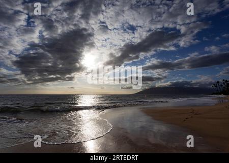Ocean surf along the shoreline of Kamaole 2 Beach at twilight with bright sunlight reflecting on the water under a cloudy sky Stock Photo