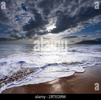 Foamy, ocean surf breaking along the shoreline of Kamaole 2 Beach at twilight with bright sunlight reflecting on the water under a cloudy sky Stock Photo