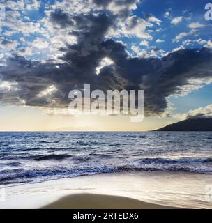 Breaking surf along the shoreline of Kamaole 2 Beach at twilight with a dramatic cloud formation in the blue sky Stock Photo