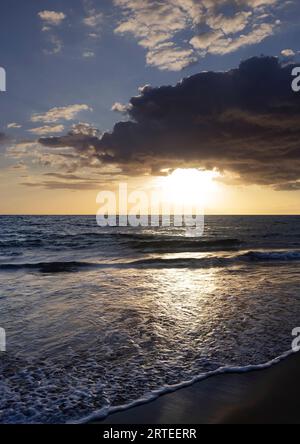 Breaking surf along the shoreline of Kamaole 2 Beach at twilight with the sun glowing behind a dramatic cloud formation in the blue sky Stock Photo